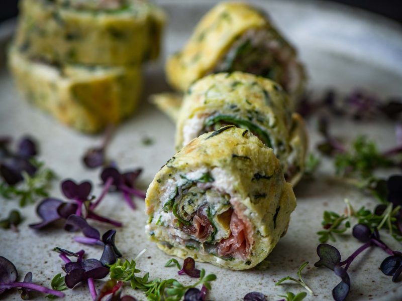 Die angeschnittene Zucchini-Ricotta-Rolle auf einem grauen Teller mit violetter und grüner Kresse nah von der Seite fotografiert.