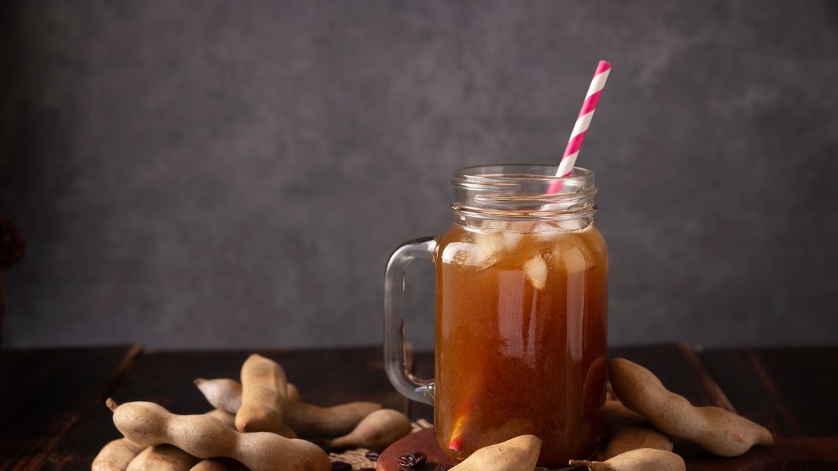 Agua de tamarindo in einem Glas mit henkel und einem gestreiften Strohhalm vor dunklem Hintergrund, daneben Tamrindenschoten.