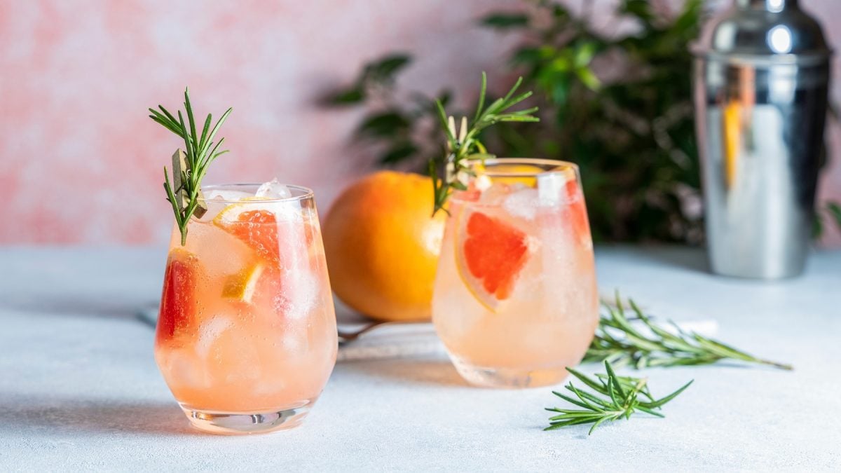 Zwei Gläser mit dem Amalfi Spritz auf einem hellen Tisch mit rötlichem Hintergrund. Alles von der Seite fotografiert und im Hintergrund noch Bar-Equipment und Pflanzen erkennbar.