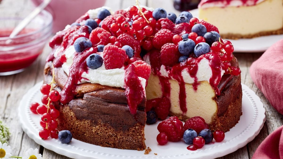 Beeren-Käsekuchen auf einem weißen Teller auf hölzernem Hintergrund. Hinten auf dem Bild ein Stück Beeren-Käsekuchen.