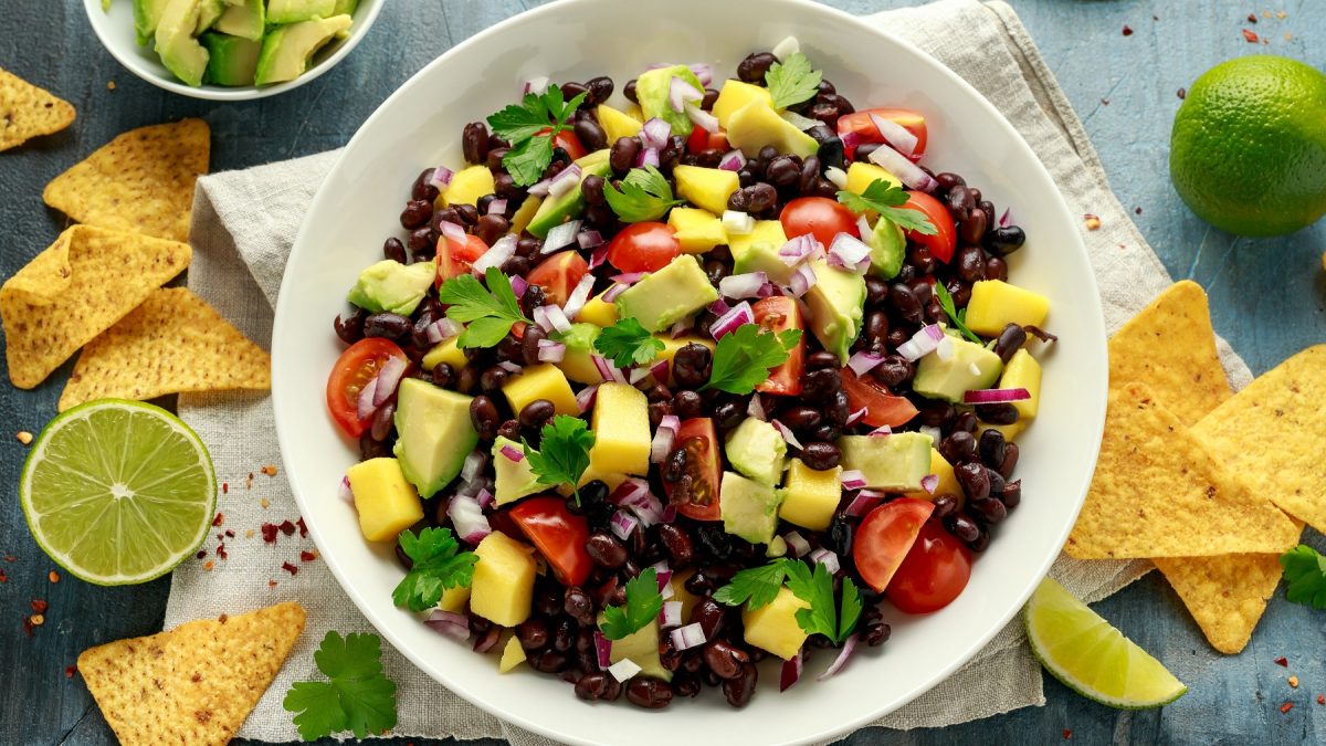 Bohnensalat mit Avocado, Mango, Tomaten, roten Zwiebeln und Mango in einer weißen Schüssel. Daneben Tortillachips und Limette.