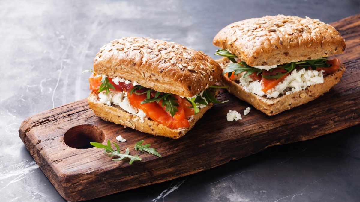 Zwei Brötchen mit Lachs und körnigem Frischkäse auf einem rustikalen Holzbrett von schräg oben fotografiert.