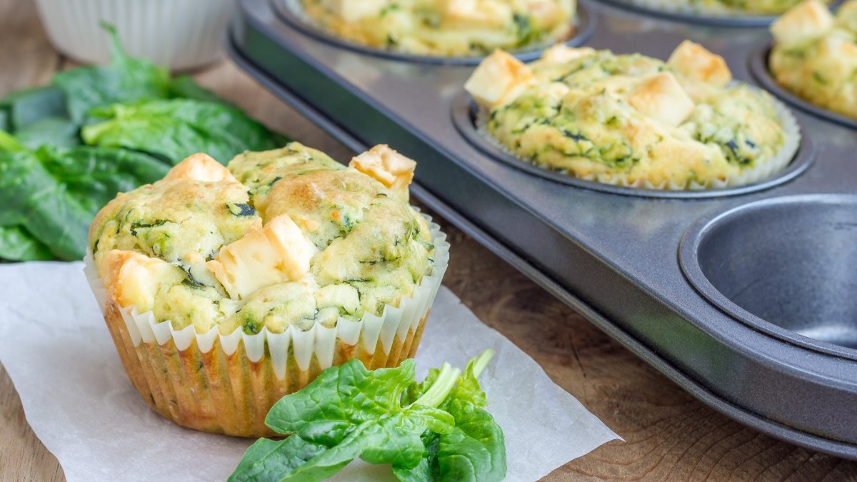 Mehrere Brokkoli-Feta-Muffins auf einem Holztisch und einige noch in der Backform dahinter. Alles von der Seite fotografiert, mit frischem Spinat.