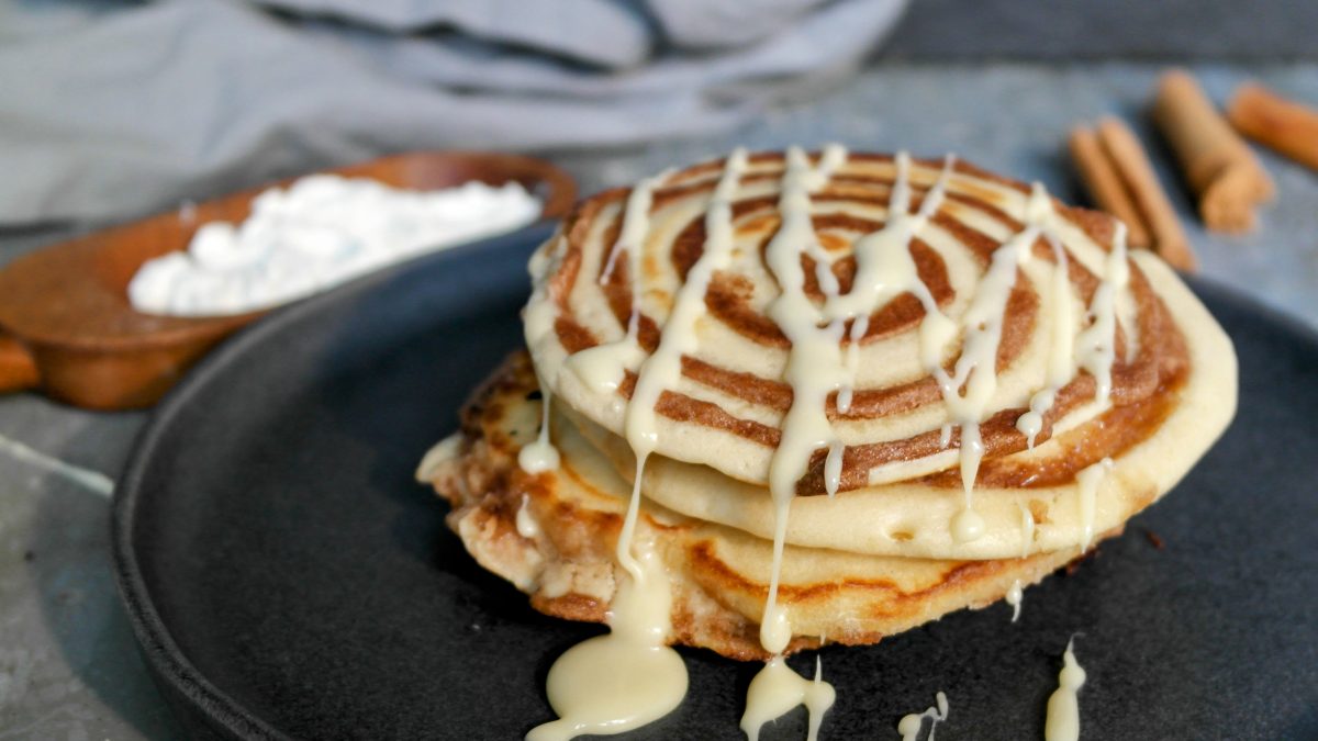 Mehrere Pfannkuchen mit Zimt-Twist auf einem dunklen Teller und grauem Tisch mit Zimt und Mehl im Hintergrund.