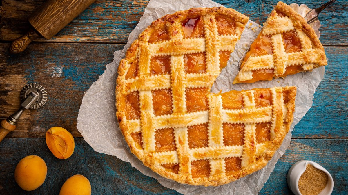 Goldbraune Crostata all'albicocca auf Backpapier mit einem Stück im Vordergrund, umgeben von frischen Aprikosen und Backutensilien.