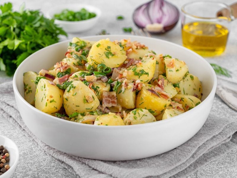Eine Schüssel mit dem perfekten Kartoffelsalat. Drum herum liegen frische Kräuter, eine halbe rote Zwiebel und andere Zutaten