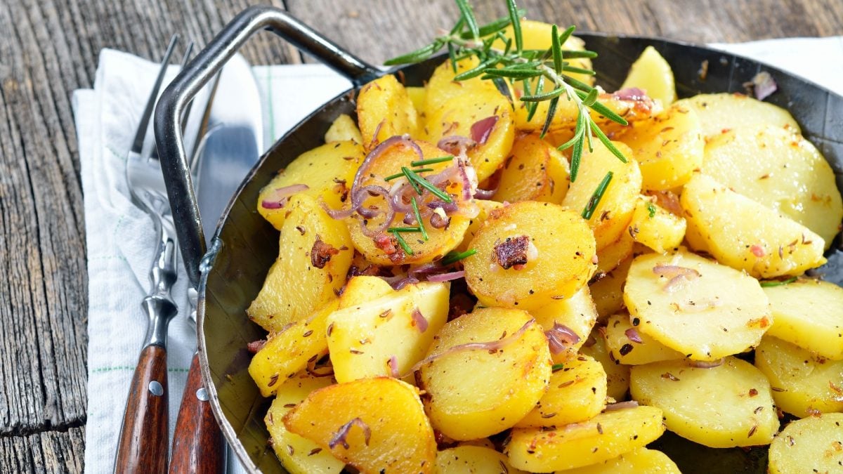 Draufsicht, schräg: Eine Pfanne mit Bratkartoffeln aus gekochten Kartoffeln. Die steht auf einem Brettchen, dass auf einem Tuch und das auf einem Holzboden.