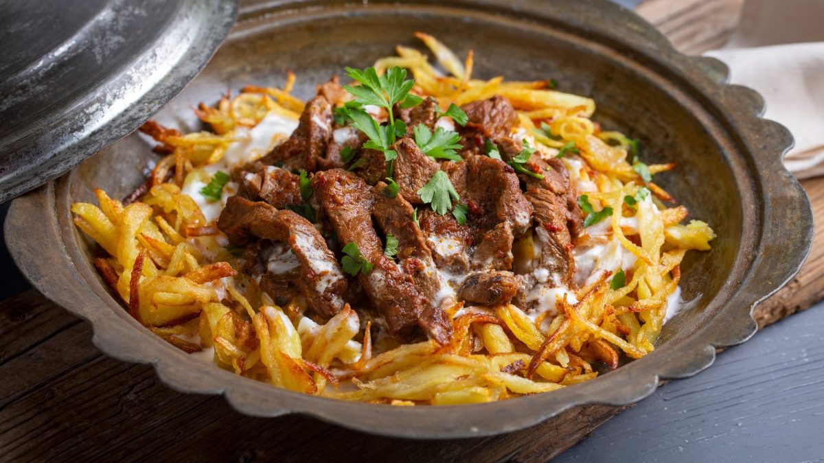 Draufsicht, schräg: Ein orientalische Metallschale Çökertme Kebabi auf einer braunen Tischdecke. Es handelt sich um ein türkisches Pommes-Rezept mit gebratenen Fleisch.