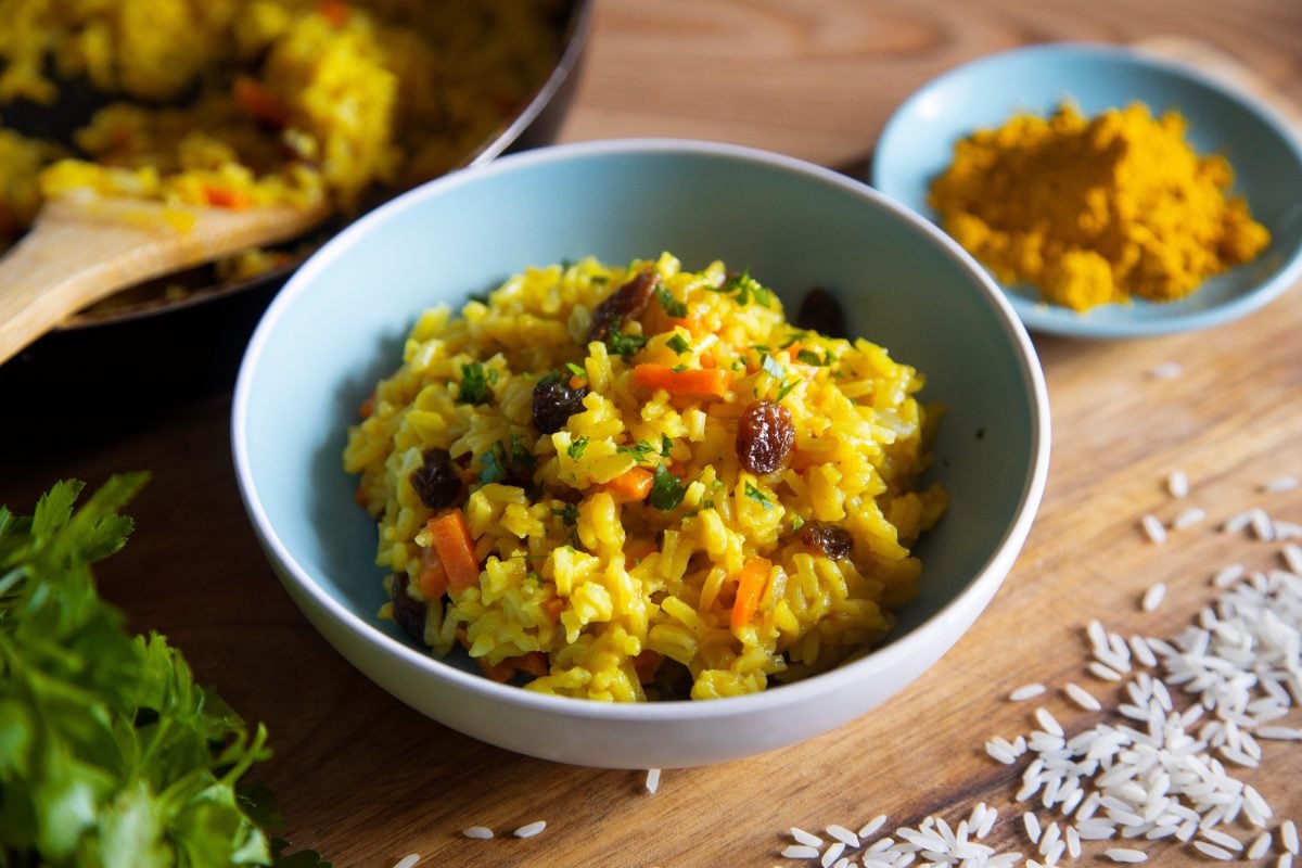 Draufsicht, schräg: eine hellblaue Schale Curry-Reis-Salat mit Rosinen auf einem Holzbrettchen. Daneben steht eine kleine hellblaue Schale mit Currypulver.