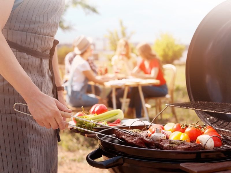 Frontal: geöffneter Grill mit verschiedenen Lebensmitteln. Ein Mensch, der sich um sie kümmert. Im Hintergrund: Eine Gruppe Menschen.