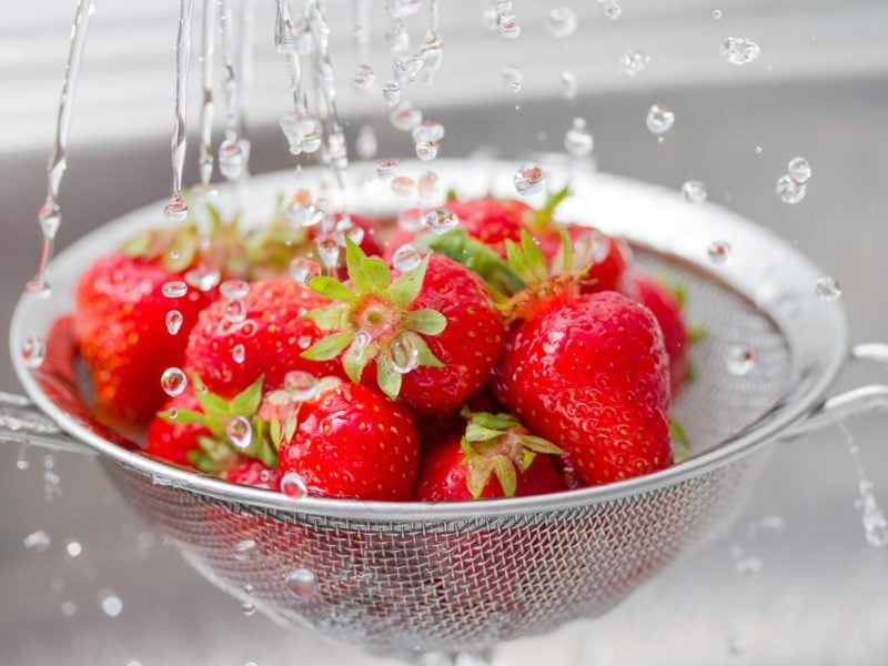 Erdbeeren waschen / Wasser aus einem Wasserhahn tropft auf Erdbeeren, die in einem Sieb aus Metall sind.