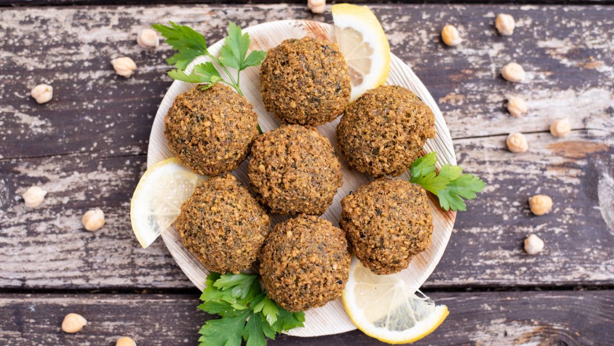 Draufsicht: Ein Teller mit Falafel aus dem Backofen steht auf einem rustikalen Holzboden. Drum herum liegen Kichererbsen.