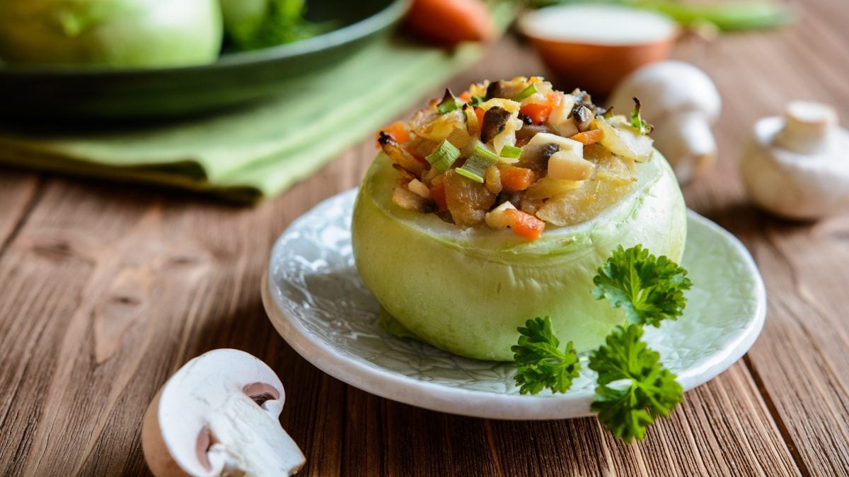 Draufsicht, schräg: eine gefüllte Kohlrabi mit Champignons steht auf einem Untergrund aus Holz. Drum herum liegen frische Pilze und Gemüse.
