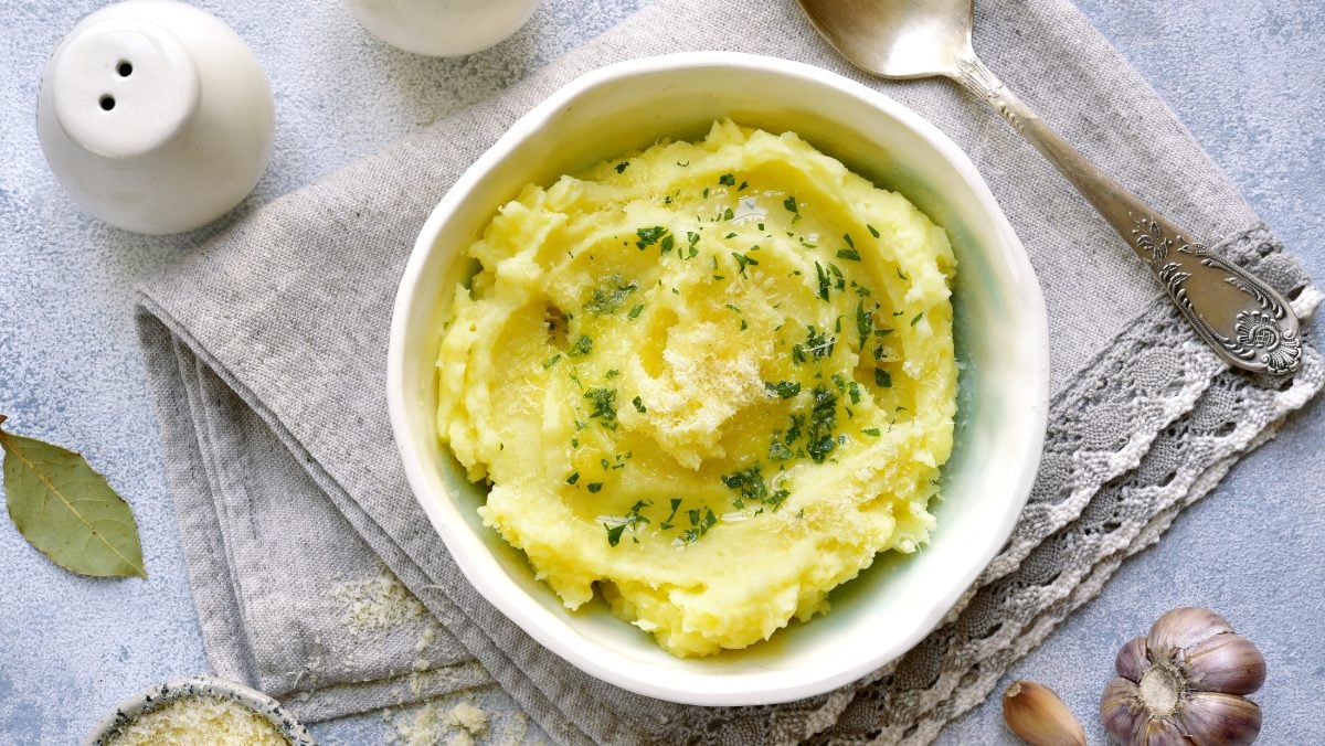 Draufsicht: eine weiße Schüssel Kartoffel-Thymian-Püree mit Käse auf einem hellblauen Untergrund aus Stoff. Daneben liegen ein silberner Löffel, ein Salzstreuer, ein Schälchen mit geriebenen Käse und ein Lorbeerblatt,