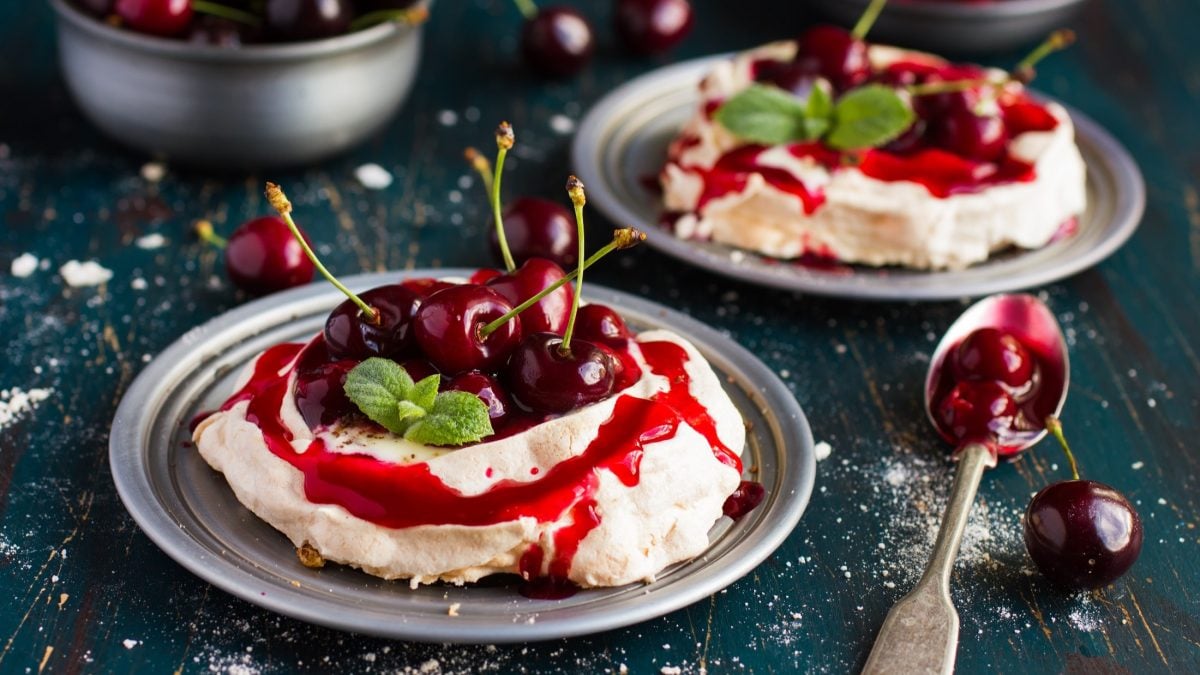 Draufsicht, schräg: Zwei Teller aus Metall mit je einer Kirsch-Pavlova. Drum herum liegen frische Kirschen und Besteck.