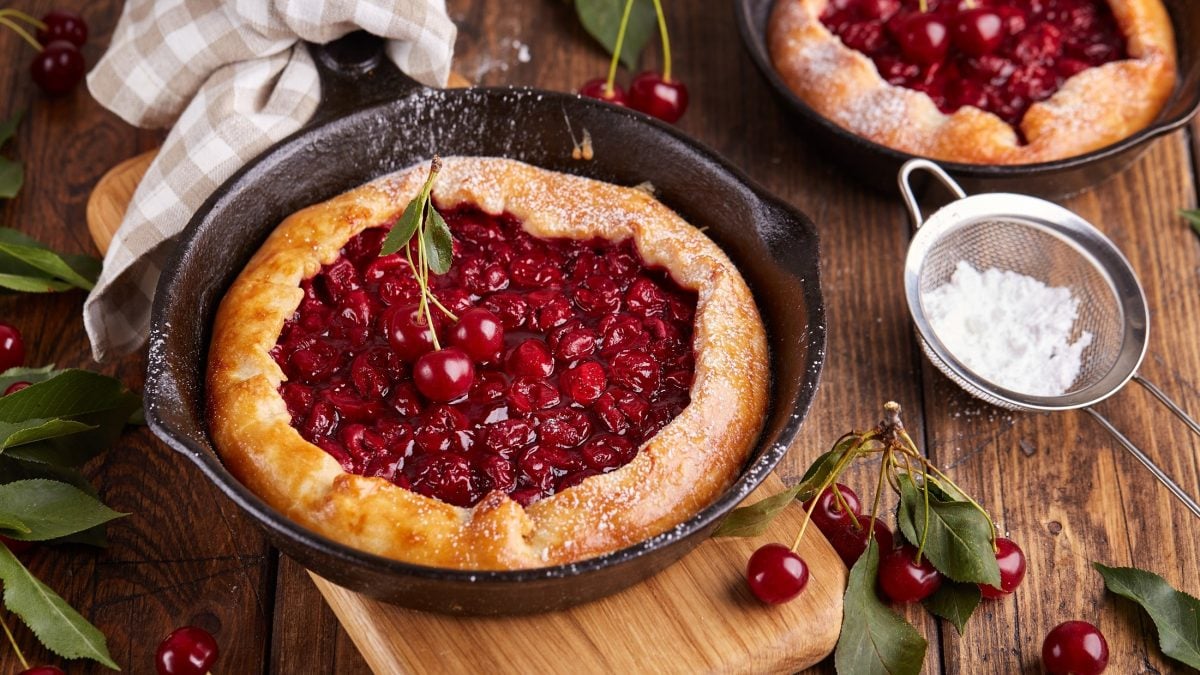 Draufsicht: Zwei Kirschen-Galette in einer ofenfesten. dunklen Form liegen auf einemr rustikalen Holzoberfläche. Drum herum sind lose Kirschen und ein weißes Tuch.