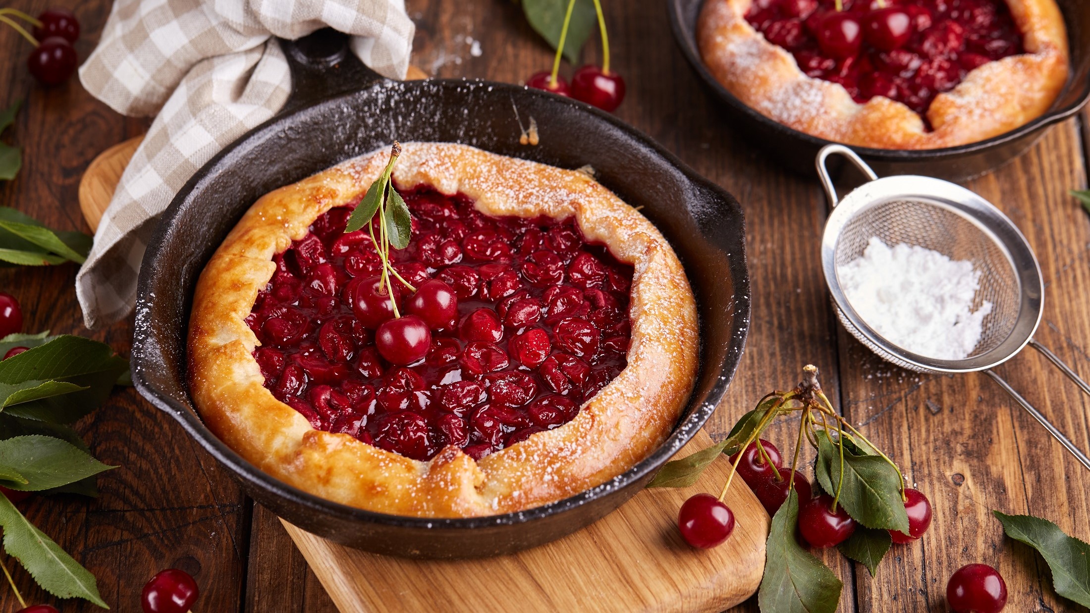 Draufsicht: Zwei Kirschen-Galette in einer ofenfesten. dunklen Form liegen auf einemr rustikalen Holzoberfläche. Drum herum sind lose Kirschen und ein weißes Tuch.