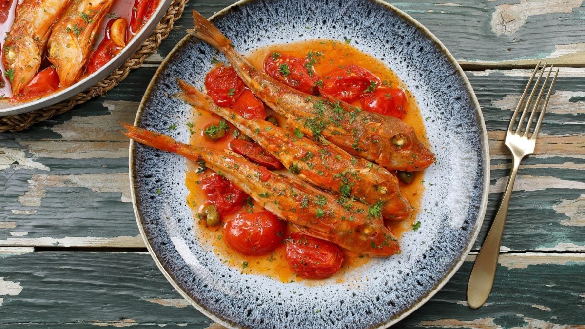 Draufsicht: Ein bläulicher, mediterran aussehender Teller Knurrhahn mit frischen Tomaten liegt auf einem bläulichem Untergrund aus Holt. Daneben liegt eine silberne Gabel.
