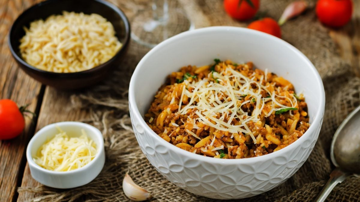 Draufsicht, schräg: Auf einem Untergrund aus Holz stehen drei Schüsseln. Eine große mit Kritharaki-Salat mit Hackfleisch. Und zwei kleine mit geriebenen Käse. Drum herum liegen einige frische Tomaten.