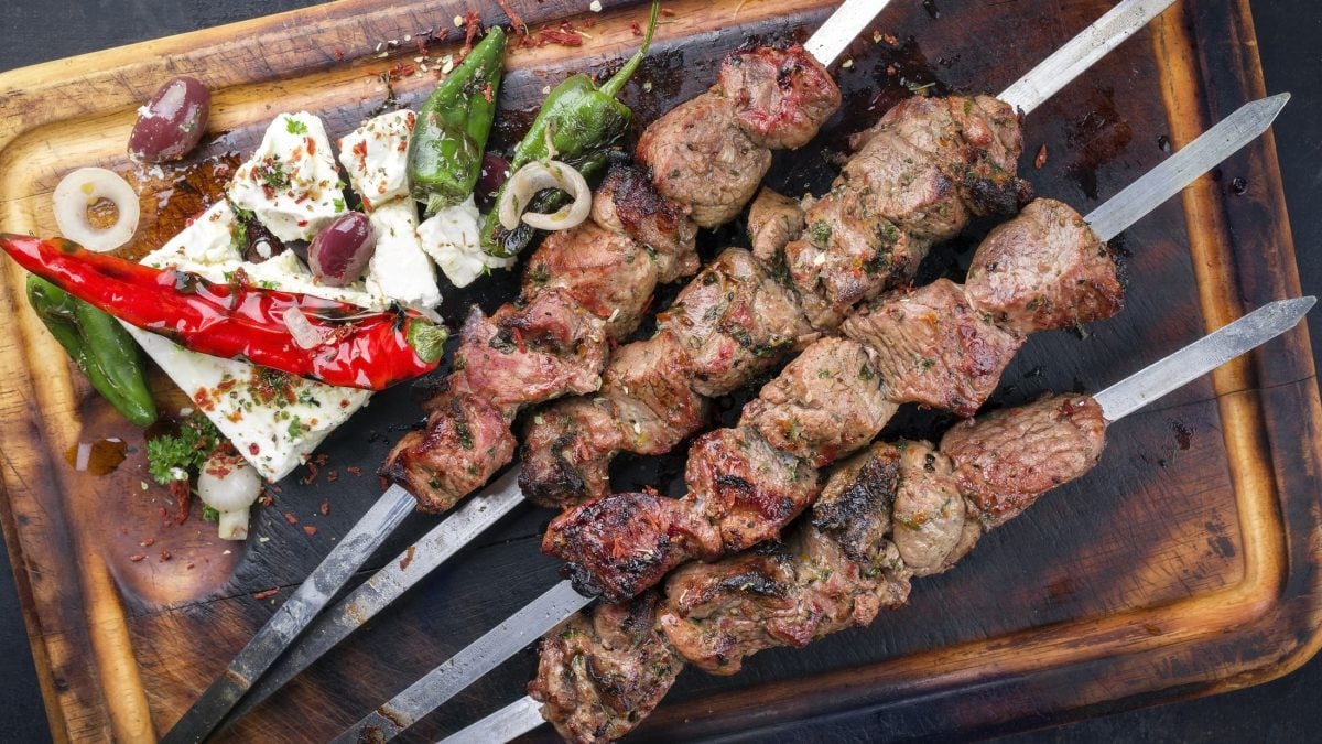 Draufsicht: Gebratene Lammspieße mit Feta und Gemüse auf einem alten, verbrannten Schneidbrett.