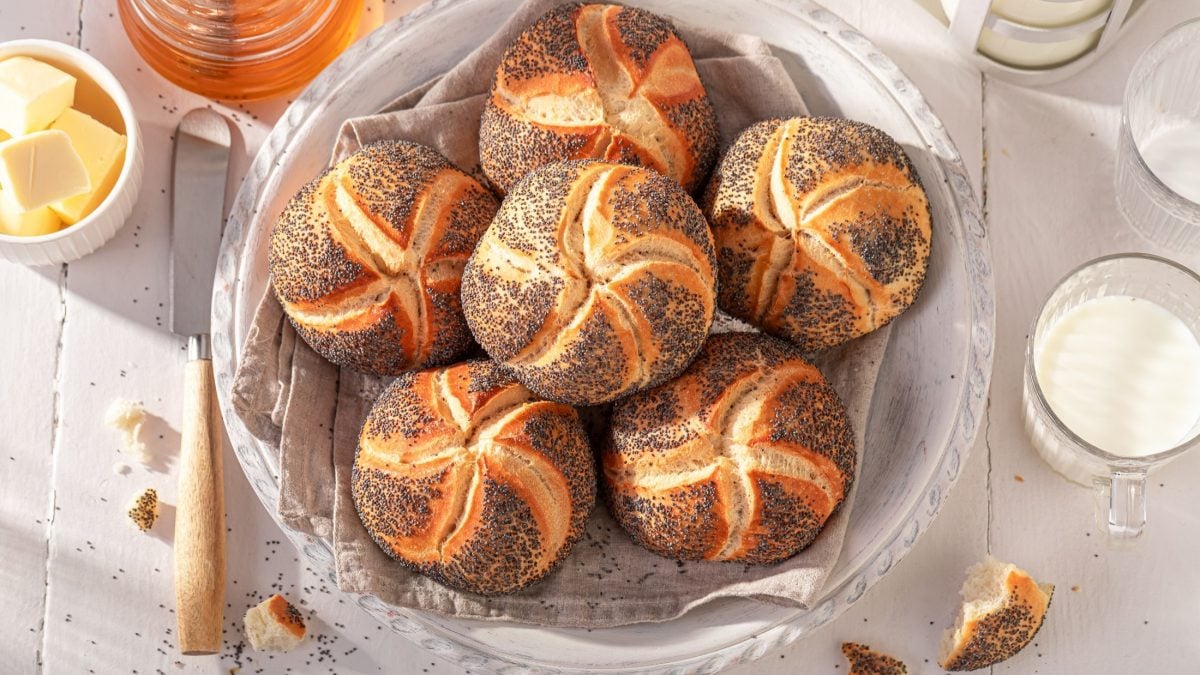 Draufsicht: Ein Frühstückstisch mit selbst gebackenen Mohnbrötchen aus Hefeteig. Drum herum sind ein weißes Schälchen mit Butter und und ein Glas Honig.