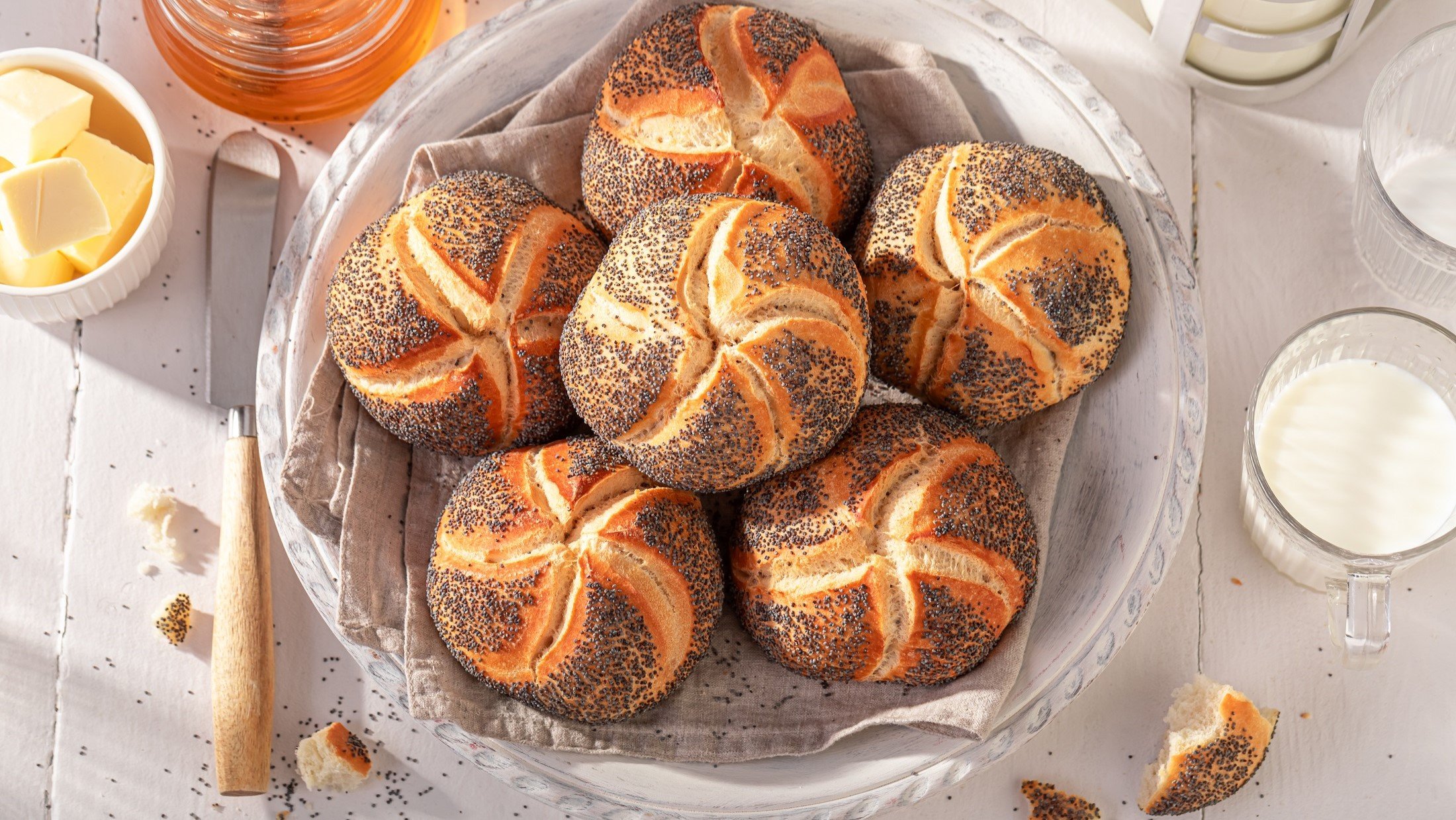 Draufsicht: Ein Frühstückstisch mit selbst gebackenen Mohnbrötchen aus Hefeteig. Drum herum sind ein weißes Schälchen mit Butter und und ein Glas Honig.