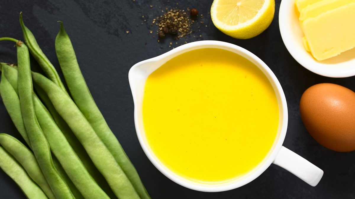 DRaufsicht: Auf einem dunklen Untergrund steht ein weißes Saucenkännchen mit einer Orangen-Hollandaise. Daneben liegen frische grübe Bohnen, eine Zitronenhälfte, ein Ei und eine weiße Schale mit Butter.