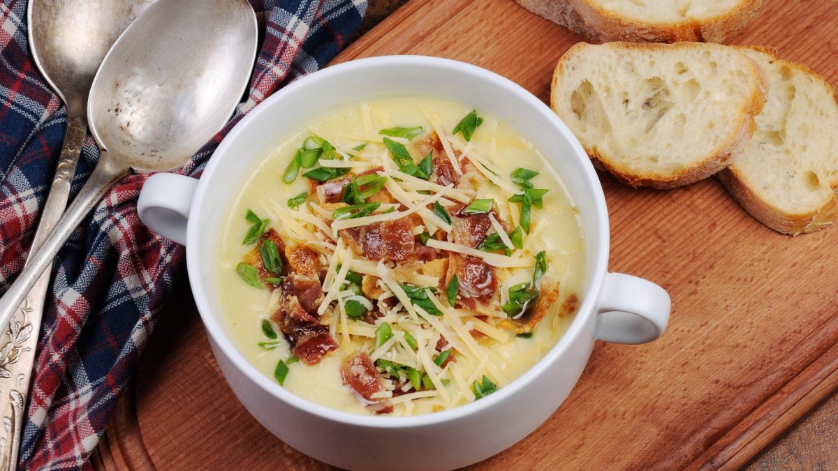 Draufsicht: Eine Pommersche Kartoffelsuppe in einer Suppentasse auf einem Untergrund aus Holz. Daneben liegen Weißbrotscheiben und zwei silberne Löffel.