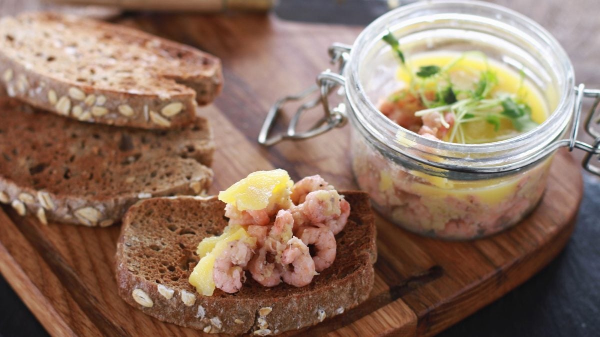 Ein Weckglas mit Potted Schrimps steht auf einem Holzbrettchen. Drum herum liegen mehrere Brotscheiben.