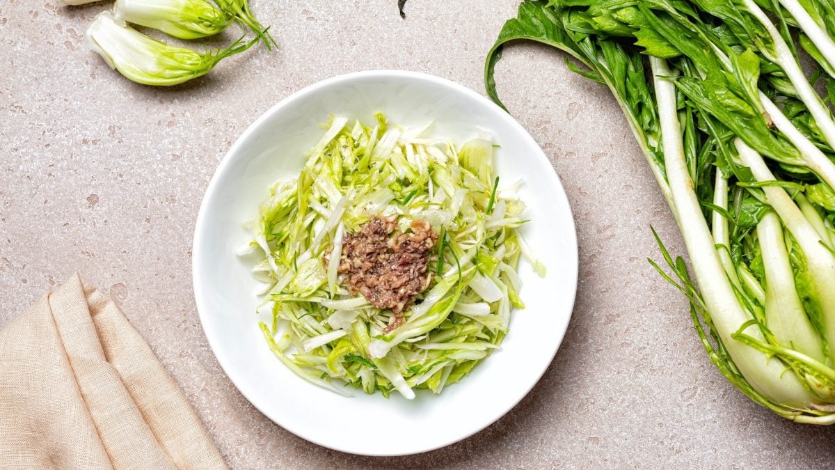 Draufsicht: Ein Teller Puntarelle-Salat mit Sardellen auf hellem Untergrund. Drum herum liegen Puntarelle-Stängel.