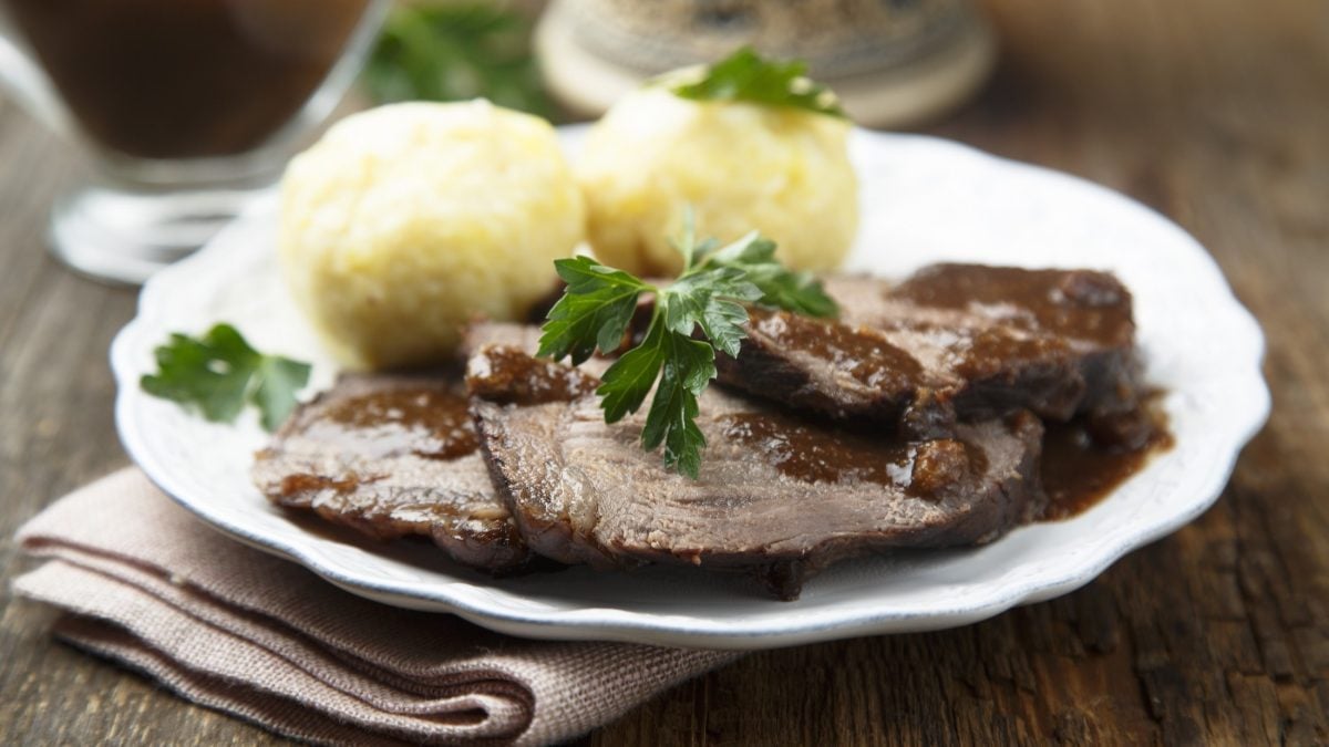 Frontal: Ein Rheinischer Sauerbraten mit zwei Kartoffelklößen auf einem weißen Teller. Das Gericht ist mit frischer Petersilie garniert.