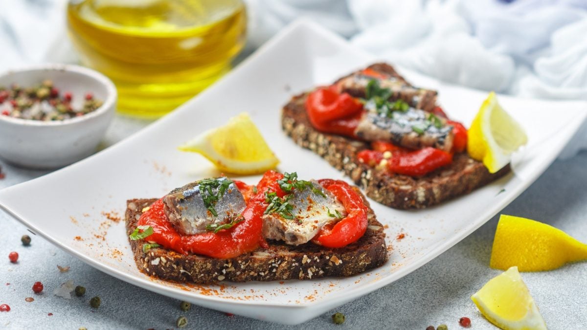 Draufsicht, schräg: Ein weißer, rechteckiger Servierteller mit zwei Sardinen-Crostini mit gerösteter Paprika. Daneben liegen mehrere Zitriónenschnitzer.