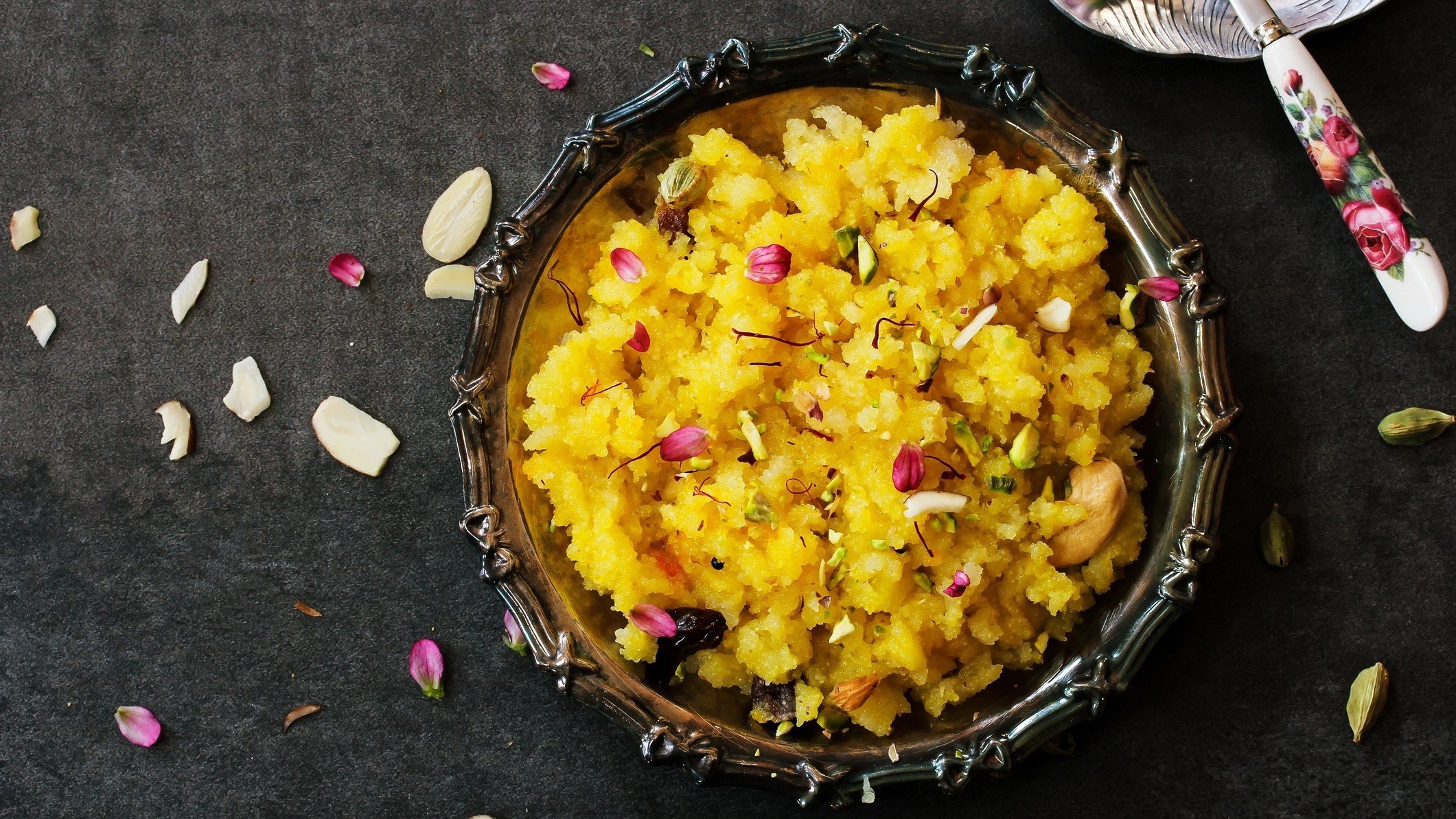 Draufsicht: Eine verzierte Schale aus Metall mit dem indischen Dessert Sooji ka halwa. Die steht auf einem dunklen Untergrund. Drum herum sind Mandeln und Rosenblätter verstreut.