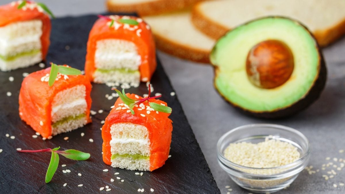 Draufsicht, schräg: Mehrere Sushi-Würfel mit Lachs liegen auf einem dunklen und einen grauen Untergrund. Daneben ist ein Schälchen mit Lachs, eine halbe Avocado und mehrere Scheiben Weißbrot.