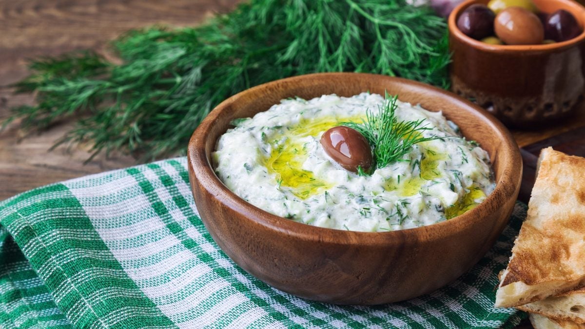 Draufsicht, schräg: Eine Tonschale mit Tsatsiki. Auf dem Dip liegen frischer Dill und ein Olive. Im Hintergrund ist eine weitere Schale mit Oliven und frischer Dill.