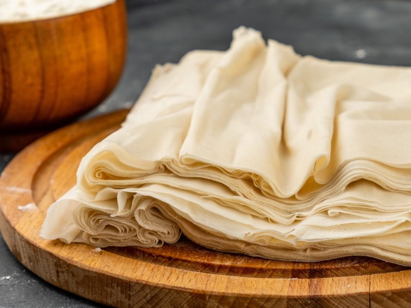 Nahaufnahme von dünnen Filoteigblättern auf einem runden Holzbrett für Filoteig-Rezepte, mit einer Holzschale im Hintergrund.