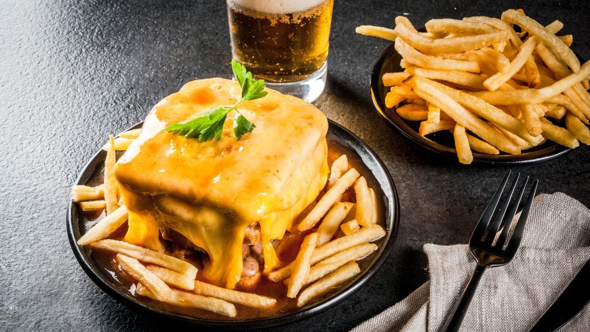 Francesinha auf einem schwarzen teller mit Pommes, daneben eine Schüssel mit Pommes und ein Glas Bier.