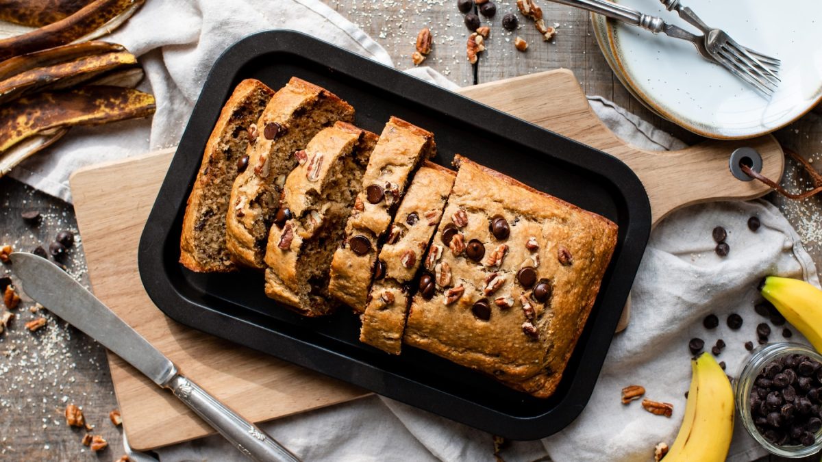 Glutenfreies Bananenbrot auf einem schwarzen Teller auf einem braunen Schneidebrett. Links ein Messer, rechts Bananen und Schokodrops.