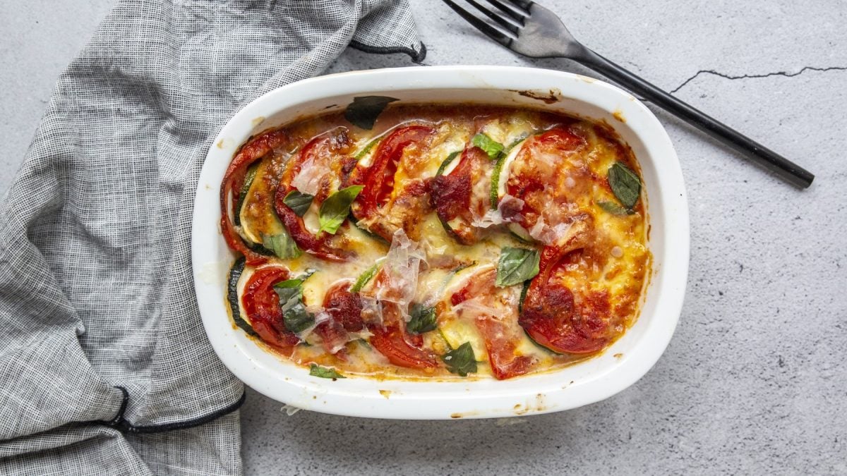 Veganes Tomaten-Kartoffel-Gratin in einer weißen Auflaufform auf grauem Hintergrund. Links ein Geschirrtuch und rechts eine Gabel.