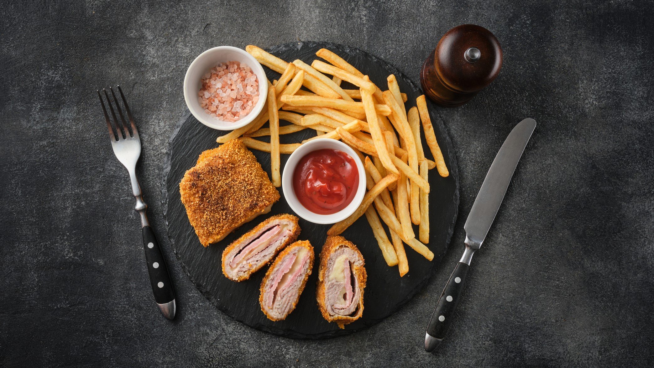 Hähnchen Cordon Bleu, aufgeschnitten, mit sichtbarer Füllung, umgeben von knusprigen Pommes, Ketchup und Gewürzen auf Steinplatte.