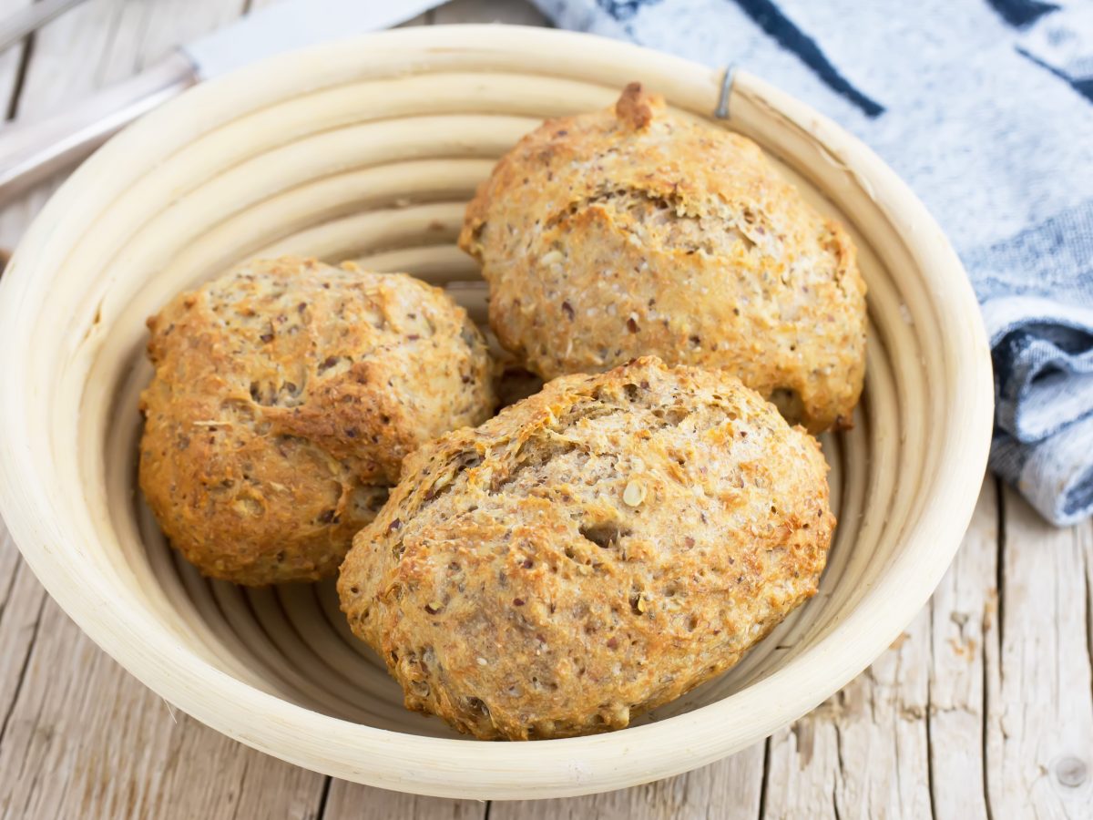 Ein heller Korb mit drei Haferflocken-Brötchen von schräg oben fotografiert.