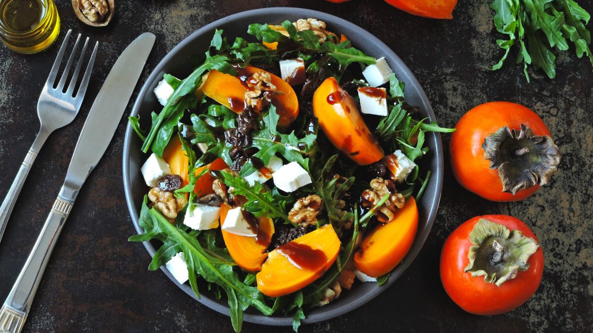 Draufsicht: Kaki-Rucola-Salat mit Feta und Dressing, daneben Kakis und Besteck, dunkler Untergrund.