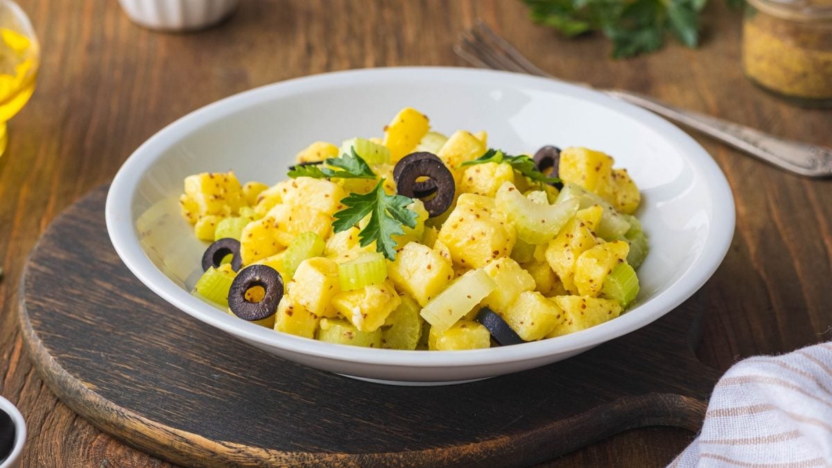 Ein weißer Teller mit dem Kartoffelsalat auf einem rustikalen Holztisch. Im Hintergrund Öl, Petersilie und Holzdeko.