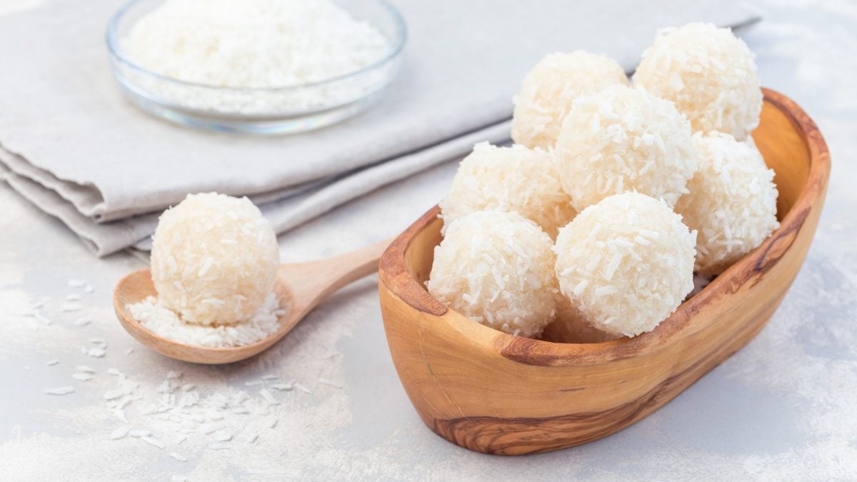 Kokos-Energy-Balls in einer hölzernen Schüssel auf hellgrauem Hintergrund. Links ein Löffel mit einem Kokos-Ball.