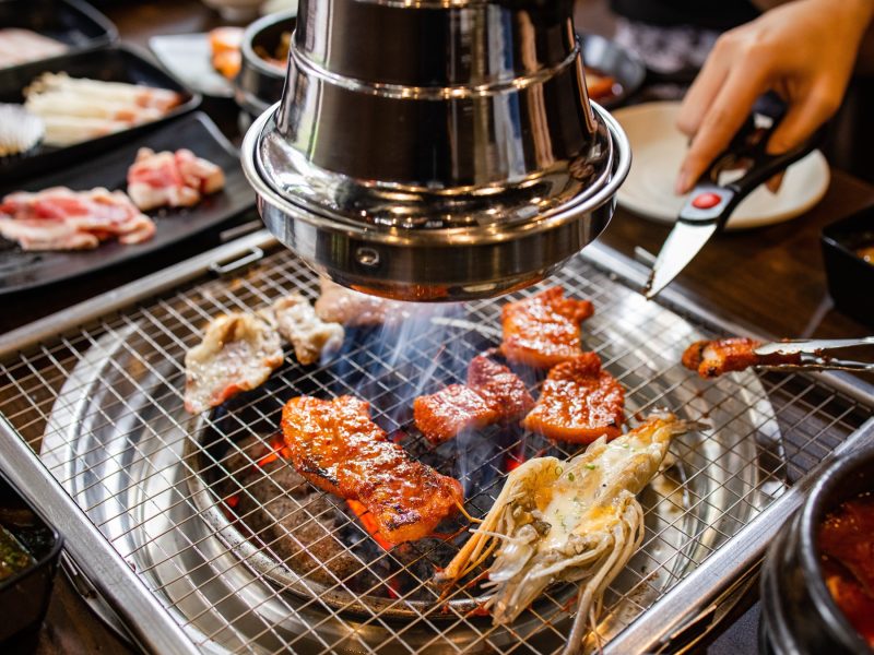Marinierte Fleischstücke und Pilze brutzeln auf einem heißen koreanischen Grill unter einer silbernen Dunstabzugshaube.