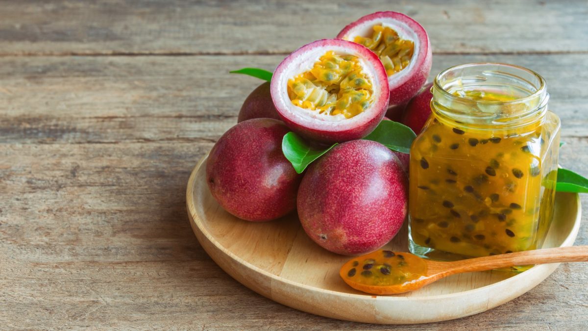 Maracuja-Marmelade in einem Glas auf einem beigen Teller auf braunem Hintergrund. Auf dem Teller sind ebenfalls Maracujas und ein Löffel.