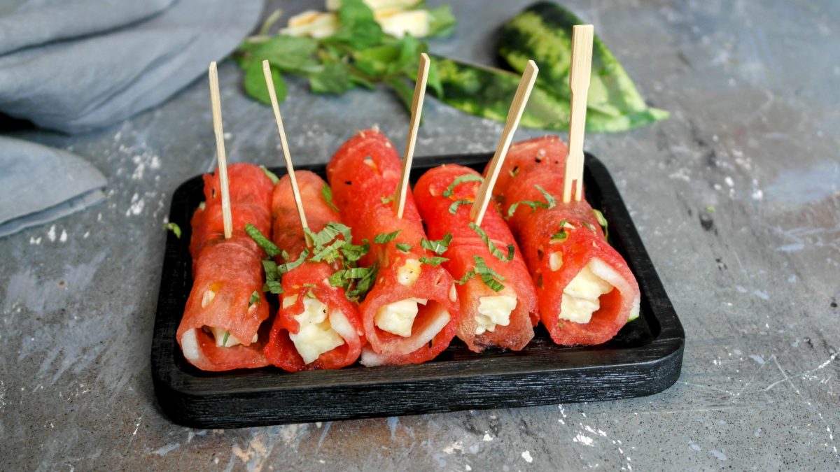Einige Melonen-Halloumi-Röllchen auf einem kleinen schwarzen Brett und Steintisch, mit Minze bestreut. Im Hintergrund unscharf Wassermelone, Minze und ein graues Tuch zu erkennen.