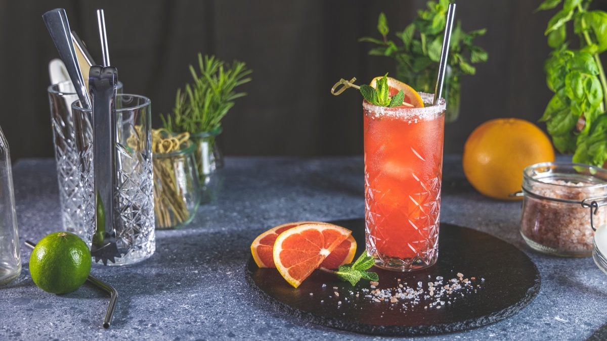 Ein Glas mit dem Mezcal Paloma auf schwarzem Teller und dunkelgrauem Untergrund. Dahinter Grapefruit, Minze und Cocktailzubehör.