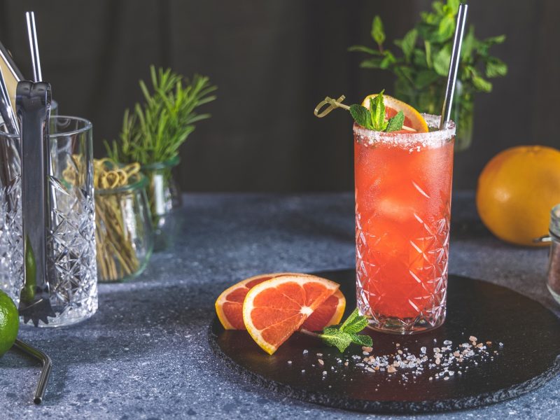 Ein Glas mit dem Mezcal Paloma auf schwarzem Teller und dunkelgrauem Untergrund. Dahinter Grapefruit, Minze und Cocktailzubehör.