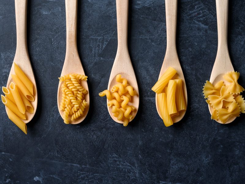 5 Holzlöffel mit verschiedenen Nudel-Sorten direkt von oben auf dunkelblauem Untergrund fotografiert.